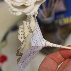 May include: A paper flower made from book pages, held on a wooden skewer. The petals are white and cream-colored, with visible printed text. The background is blurred, with hints of blue and yellow.