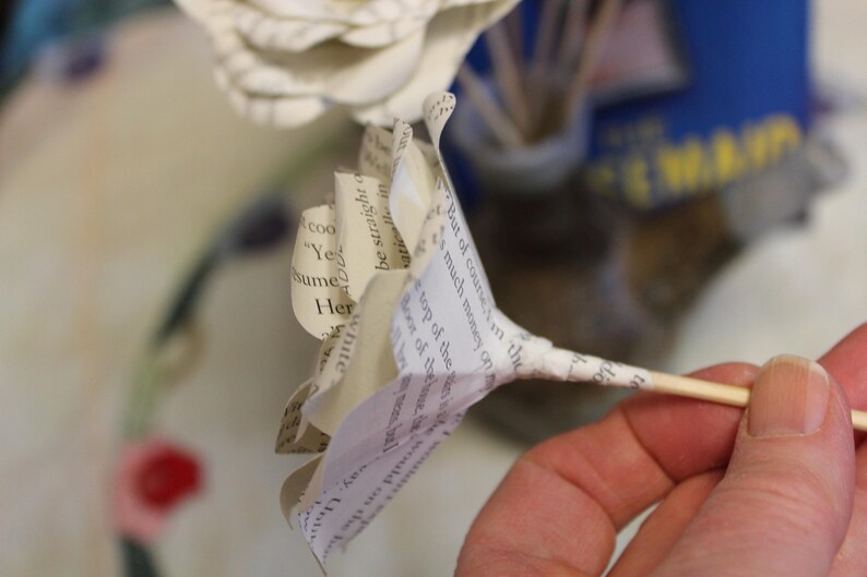 May include: A paper flower made from book pages, held on a wooden skewer. The petals are white and cream-colored, with visible printed text. The background is blurred, with hints of blue and yellow.