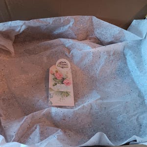 May include: A cardboard box containing white tissue paper with glitter. A decorative tag featuring pink roses and the word "Friends" is visible.