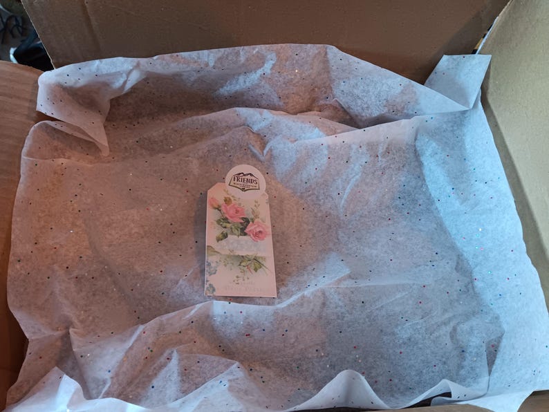May include: A cardboard box containing white tissue paper with glitter. A decorative tag featuring pink roses and the word "Friends" is visible.