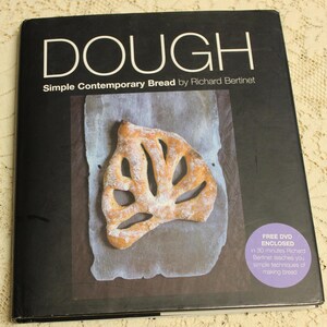 May include: A black book titled "DOUGH Simple Contemporary Bread by Richard Bertinet" with a photo of a loaf of bread on the cover. A purple circle with white text says "FREE DVD ENCLOSED in 30 minutes Richard Bertinet teaches you simple techniques of making bread".