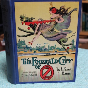 May include: A vintage hardcover book titled "The Emerald City of Oz" by L. Frank Baum. The cover features a colorful illustration of a woman riding a dragon-like creature over a city. The book has a blue cover and spine.