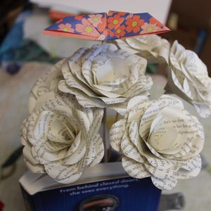 May include: A bouquet of paper roses made from book pages, with a folded paper origami butterfly above. The roses are white with visible text, and the butterfly has a blue and orange floral pattern. The bouquet is in a blue container.