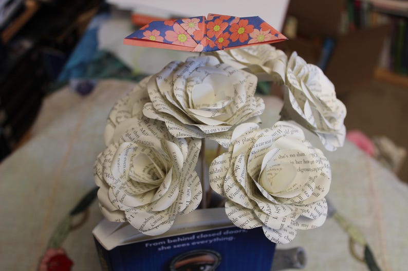May include: A bouquet of paper roses made from book pages, with a folded paper origami butterfly above. The roses are white with visible text, and the butterfly has a blue and orange floral pattern. The bouquet is in a blue container.