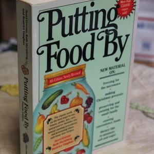 May include: A hardcover book titled "Putting Food By" with a light green cover. The cover features a jar illustration with various fruits and vegetables. Text includes "4th Edition, Newly Revised" and "More than 550,000 copies sold."