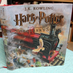 May include: A hardcover book titled "Harry Potter and the Sorcerer's Stone" by J.K. Rowling. The cover art features a red steam train, an owl, and the title in gold lettering. The book is illustrated by Jim Kay.