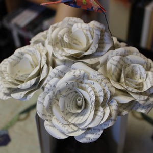 May include: A bouquet of paper roses crafted from book pages. The white roses, displaying visible text, are arranged in a dark vase. A small, colourful bird decoration is attached to the arrangement.