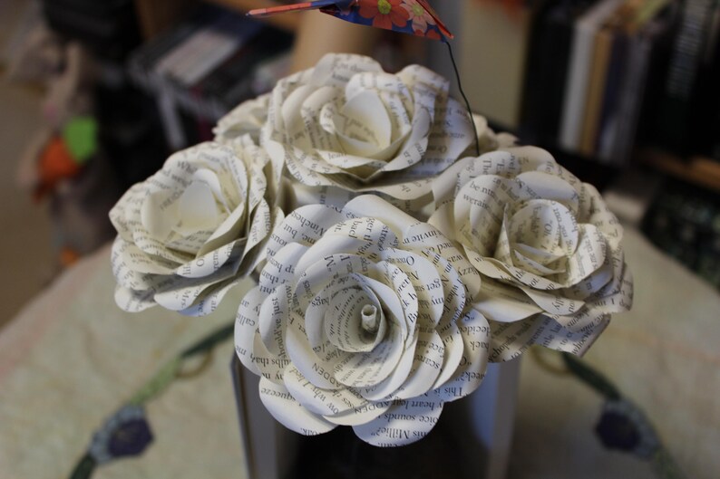 May include: A bouquet of paper roses crafted from book pages. The white roses, displaying visible text, are arranged in a dark vase. A small, colourful bird decoration is attached to the arrangement.