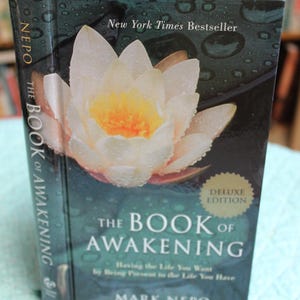 Może przedstawiać: Książka w twardej oprawie zatytułowana "The Book of Awakening" autorstwa Marka Nepo. Okładka przedstawia biało-ż&oacute;łty kwiat z kroplami wody, widoczny jest tekst "New York Times Bestseller". Obecna jest r&oacute;wnież naklejka "Deluxe Edition".