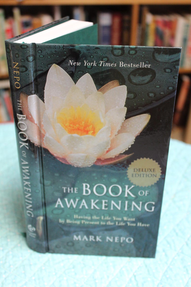 Może przedstawiać: Książka w twardej oprawie zatytułowana "The Book of Awakening" autorstwa Marka Nepo. Okładka przedstawia biało-ż&oacute;łty kwiat z kroplami wody, widoczny jest tekst "New York Times Bestseller". Obecna jest r&oacute;wnież naklejka "Deluxe Edition".