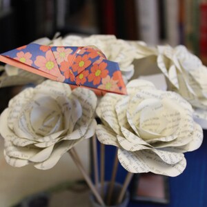 May include: A bouquet of paper flowers, featuring white roses crafted from book pages and a folded origami crane. The crane has a floral pattern in orange, pink, and yellow on a blue background.