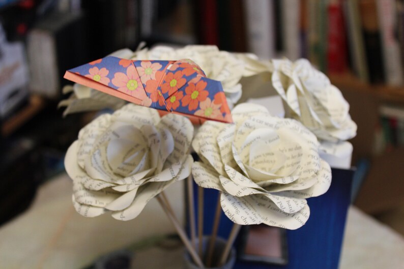 May include: A bouquet of paper flowers, featuring white roses crafted from book pages and a folded origami crane. The crane has a floral pattern in orange, pink, and yellow on a blue background.