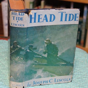 May include: Vintage hardcover book titled "Head Tide" by Joseph C. Lincoln. The cover features a blue and white design with a painting of men in a boat on the water. The book's spine is worn, showing signs of age.