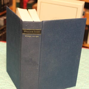 May include: A dark blue hardcover book titled William James Writings, 1902-1910 with gold lettering on the spine. The book is positioned upright in front of a white slipcase, showing a small gold emblem of an open book with stars at the base.