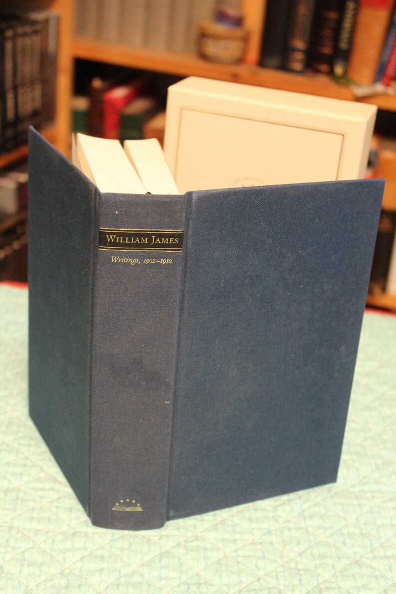 May include: A dark blue hardcover book titled William James Writings, 1902-1910 with gold lettering on the spine. The book is positioned upright in front of a white slipcase, showing a small gold emblem of an open book with stars at the base.