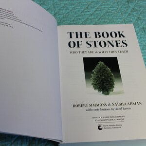 May include: Open book titled "THE BOOK OF STONES" with a green stone image. The book's authors are Robert Simmons and Naisha Ahsian. The book is open on a turquoise quilted surface.
