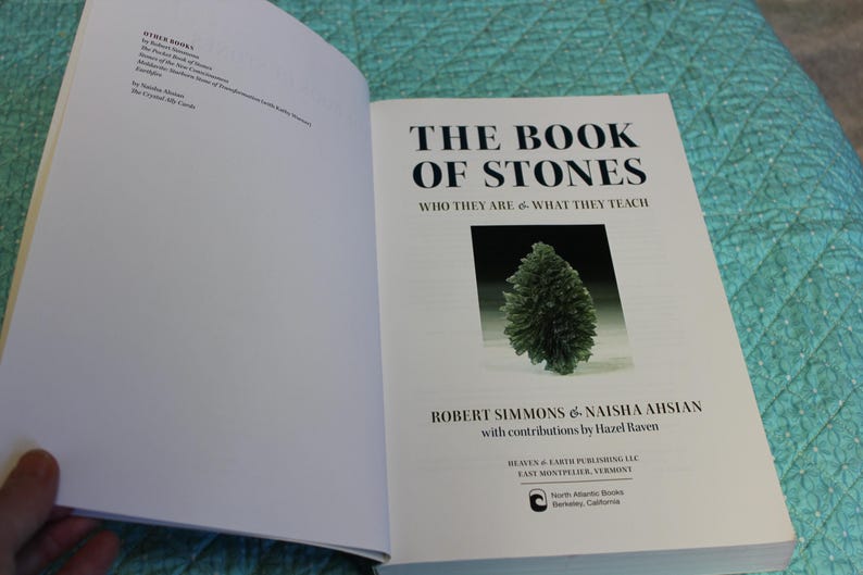 May include: Open book titled "THE BOOK OF STONES" with a green stone image. The book's authors are Robert Simmons and Naisha Ahsian. The book is open on a turquoise quilted surface.
