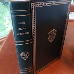 May include: A dark green hardcover book titled Aesop, Grimm, Andersen from The Harvard Classics Deluxe Edition. The spine features gold lettering and a shield emblem with the text VERITAS. The front cover has a gold border and shield.