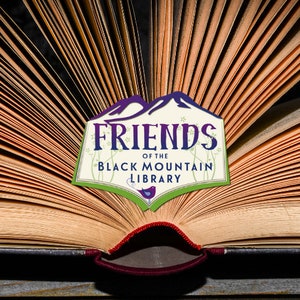 May include: An open book with aged, yellowed pages, fanned out to display the text. A logo reading "FRIENDS of the BLACK MOUNTAIN LIBRARY" is centered on the pages. The logo features a purple mountain range and green accents.