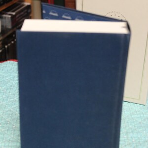 May include: A dark blue hardcover book with white pages standing upright. The book features a dark blue interior lining with a repeating white book icon pattern. Behind the book is a cream-colored slipcase with a thin gold border and text.