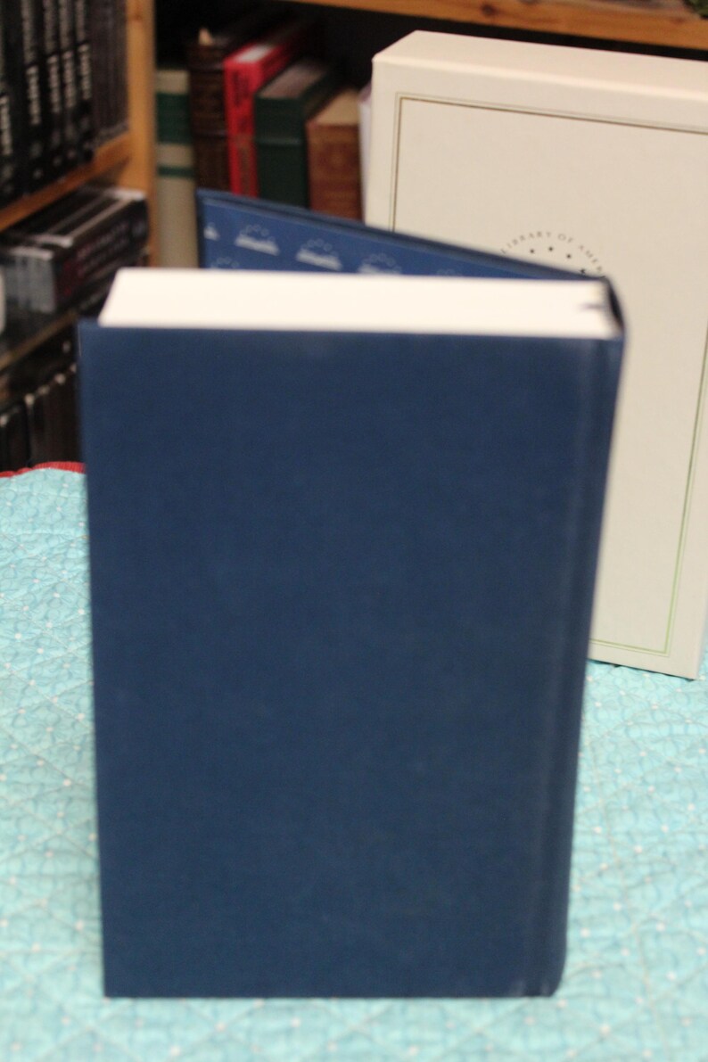 May include: A dark blue hardcover book with white pages standing upright. The book features a dark blue interior lining with a repeating white book icon pattern. Behind the book is a cream-colored slipcase with a thin gold border and text.
