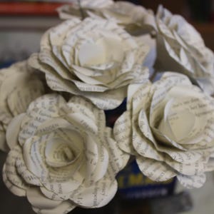 May include: A close-up of a bouquet of paper roses crafted from book pages. The cream-coloured flowers, with visible text, are arranged in a blue container. The layered petals give a realistic floral look.