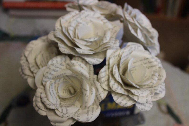May include: A close-up of a bouquet of paper roses crafted from book pages. The cream-coloured flowers, with visible text, are arranged in a blue container. The layered petals give a realistic floral look.