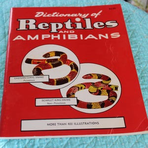 May include: A red book titled "Dictionary of Reptiles and Amphibians" with illustrations of snakes. The cover features the text "Eastern Coral Snake" and "Scarlet King Snake". The book has over 400 illustrations.