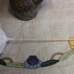 May include: A paper flower with a wooden stem. The flower is crafted from book pages, featuring a white petal design. The stem is a thin, light brown wooden dowel. The flower rests on a floral patterned surface.