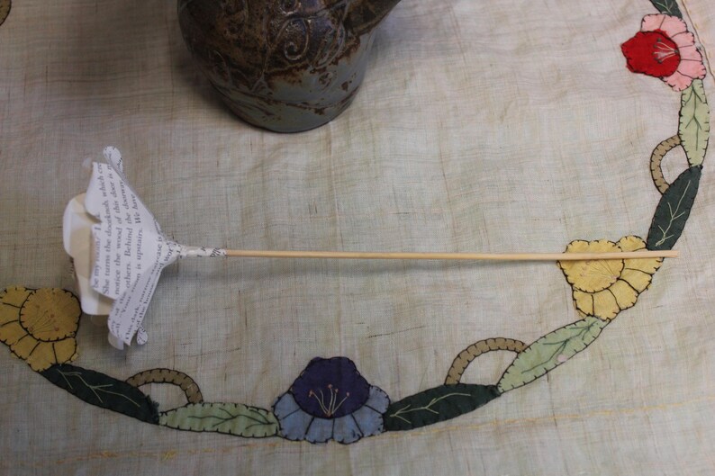 May include: A paper flower with a wooden stem. The flower is crafted from book pages, featuring a white petal design. The stem is a thin, light brown wooden dowel. The flower rests on a floral patterned surface.
