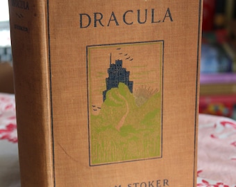 Puede incluir: Un libro antiguo de tapa dura titulado "Drácula" de Bram Stoker. La portada presenta un fondo marrón con una ilustración en blanco y negro de un castillo en una colina.