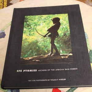 May include: A hardcover book titled "Efe Pygmies: Archers of the African Rain Forest." The cover features a color photograph of a person holding a bow and arrow in a forest setting. Text includes the author's name.