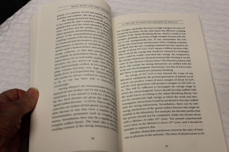 May include: A close-up of a book open to a page with text about black holes and baby universes. The text discusses the strong and weak interactions of particles and the energy required to unify them.