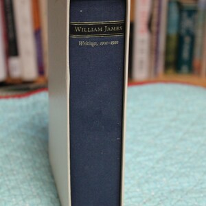 May include: A navy blue hardcover book titled William James Writings, 1902-1910, encased in a white slipcase. The spine features gold lettering and a small gold emblem at the bottom. A thin navy blue ribbon bookmark extends from the bottom edge.