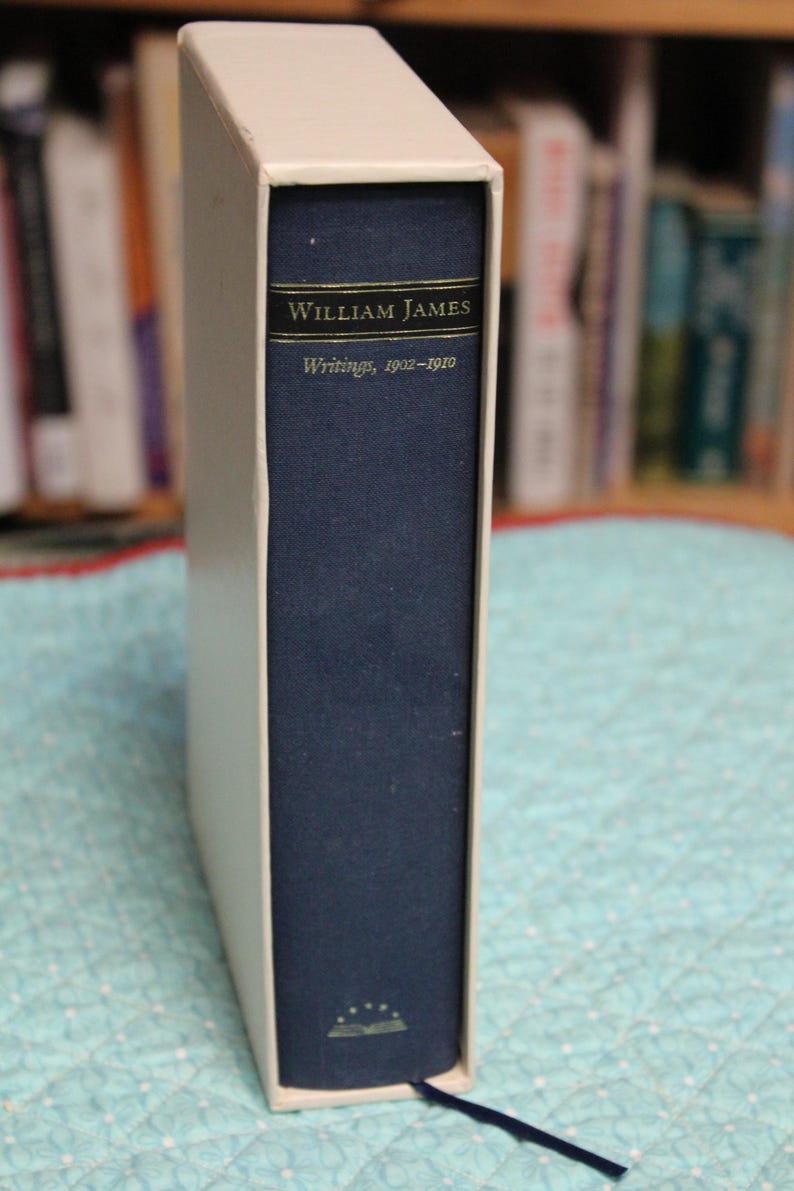 May include: A navy blue hardcover book titled William James Writings, 1902-1910, encased in a white slipcase. The spine features gold lettering and a small gold emblem at the bottom. A thin navy blue ribbon bookmark extends from the bottom edge.