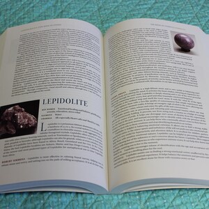 May include: An open book with text and images. One page features a photo of a purple stone and the other a photo of a cluster of dark red-brown crystals. The text includes the word "LEPIDOLITE". The book is on a teal surface.