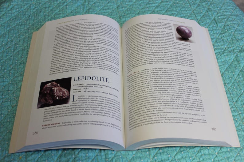 May include: An open book with text and images. One page features a photo of a purple stone and the other a photo of a cluster of dark red-brown crystals. The text includes the word "LEPIDOLITE". The book is on a teal surface.