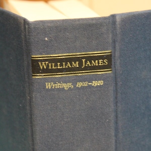 May include: A dark blue hardcover book spine featuring a black rectangular label with gold lettering that reads William James and Writings, 1902-1910. The book has a textured fabric cover and is positioned upright with white pages visible at the top.