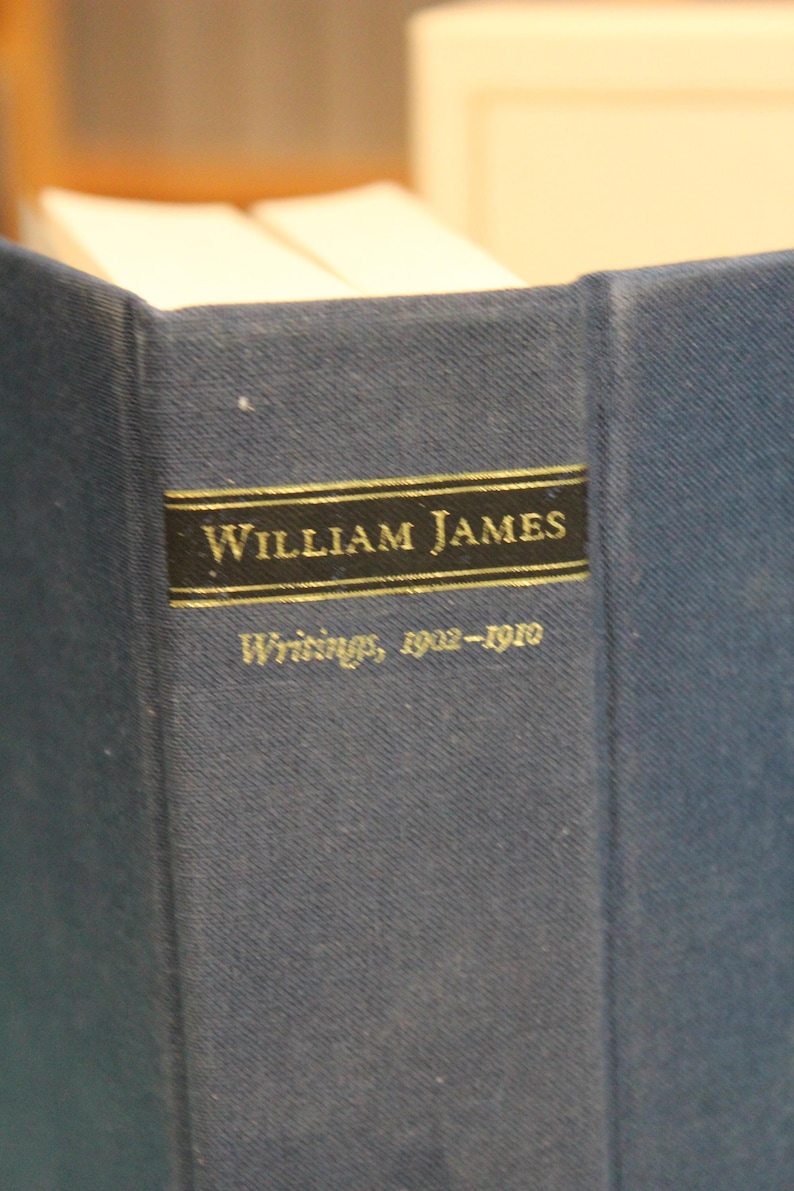 May include: A dark blue hardcover book spine featuring a black rectangular label with gold lettering that reads William James and Writings, 1902-1910. The book has a textured fabric cover and is positioned upright with white pages visible at the top.