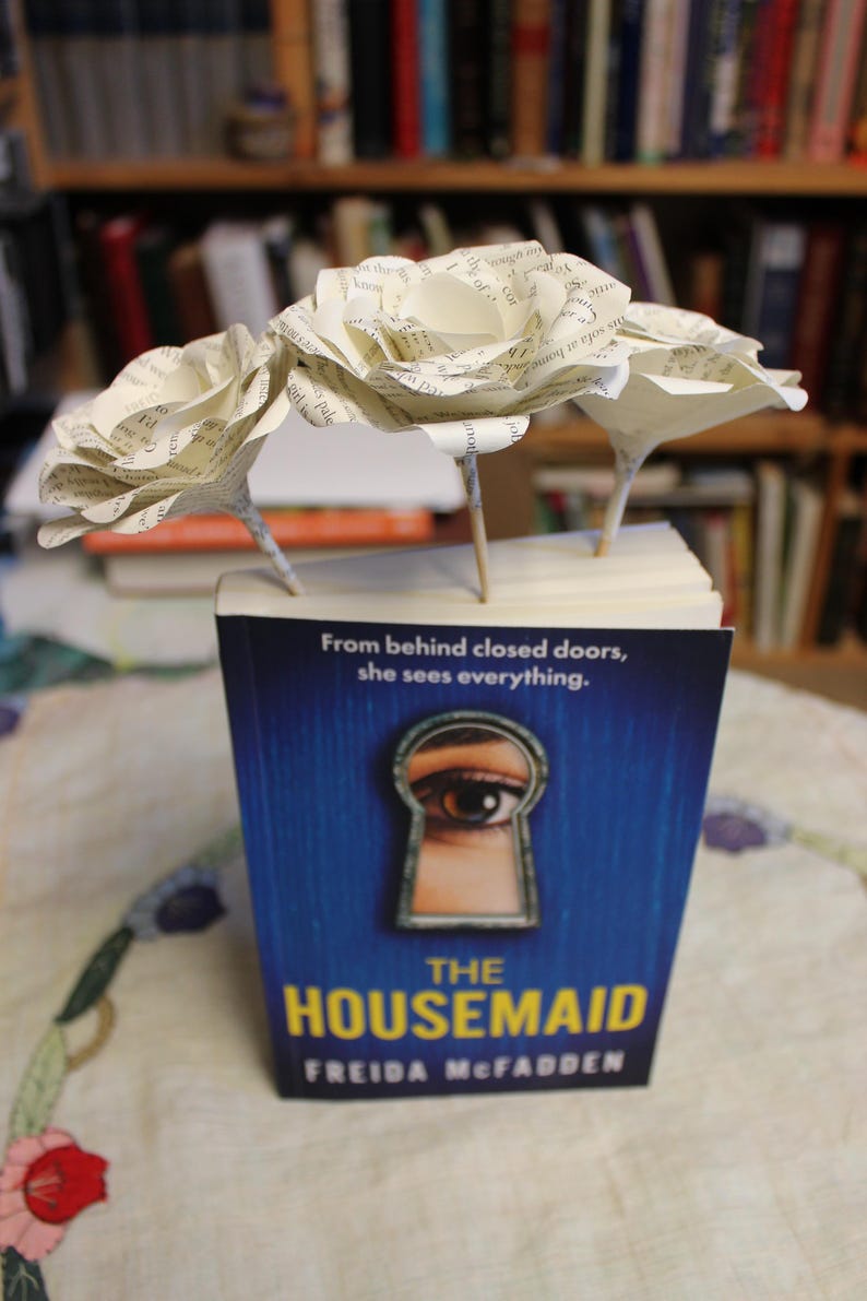 May include: A copy of "The Housemaid" with a blue cover and a keyhole design, adorned with three paper flowers. The flowers are crafted from book pages and have wooden stems. The book rests on a table.