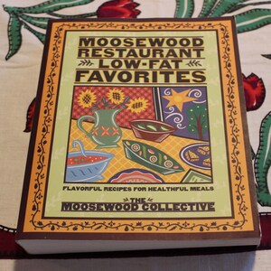 May include: A brown cookbook with a yellow and orange border. The cover features a colorful illustration of a restaurant scene with the text "MOOSEWOOD RESTAURANT LOW-FAT FAVORITES THE MOOSEWOOD COLLECTIVE".