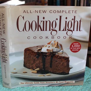 May include: A cookbook titled "All-New Complete Cooking Light Cookbook" with a slice of chocolate cake on the cover. The cake is topped with whipped cream and almonds. The book cover also includes the text "Over 1,000 of our best recipes."