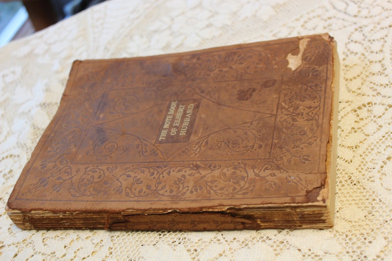 May include: A worn brown leather-bound book with the title "The Note Book of Elbert Hubbard" embossed in gold lettering. The cover features an intricate floral design.