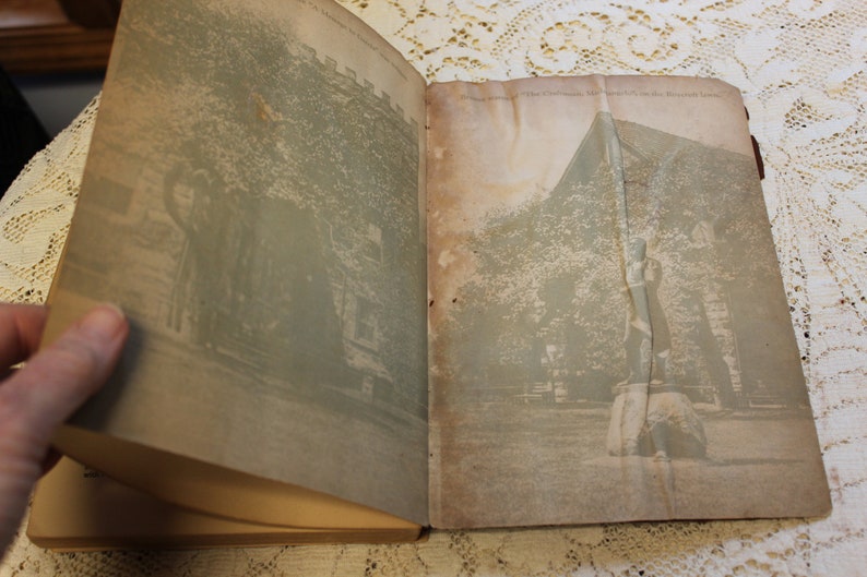 May include: An open book with two black and white photographs. The left page shows a building with trees in front of it. The right page shows a statue of a person standing on a rock with trees in the background.