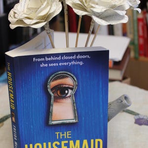 May include: A copy of "THE HOUSEMAID" by Freida McFadden, with a blue cover and an eye in a keyhole design. Paper flowers are arranged in the book. The text "From behind closed doors, she sees everything" is visible.