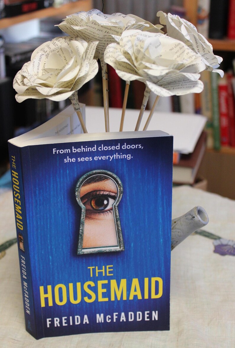 May include: A copy of "THE HOUSEMAID" by Freida McFadden, with a blue cover and an eye in a keyhole design. Paper flowers are arranged in the book. The text "From behind closed doors, she sees everything" is visible.
