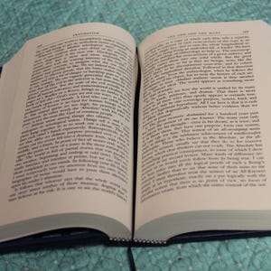 May include: An open hardcover book with cream-colored pages filled with black printed text. The book features a dark blue cover with a subtle white pattern along the edges and a thin dark blue ribbon bookmark extending from the bottom spine.