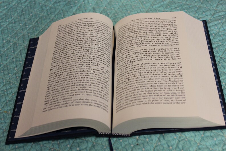 May include: An open hardcover book with cream-colored pages filled with black printed text. The book features a dark blue cover with a subtle white pattern along the edges and a thin dark blue ribbon bookmark extending from the bottom spine.