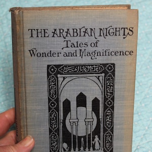 May include: A vintage hardcover book titled "The Arabian Nights Tales of Wonder and Magnificence." The cover is a light gray with black text and an ornate illustration. The book is held by a hand, suggesting its size and age.