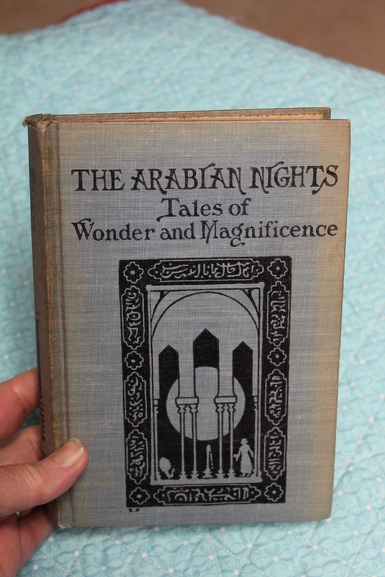 May include: A vintage hardcover book titled "The Arabian Nights Tales of Wonder and Magnificence." The cover is a light gray with black text and an ornate illustration. The book is held by a hand, suggesting its size and age.